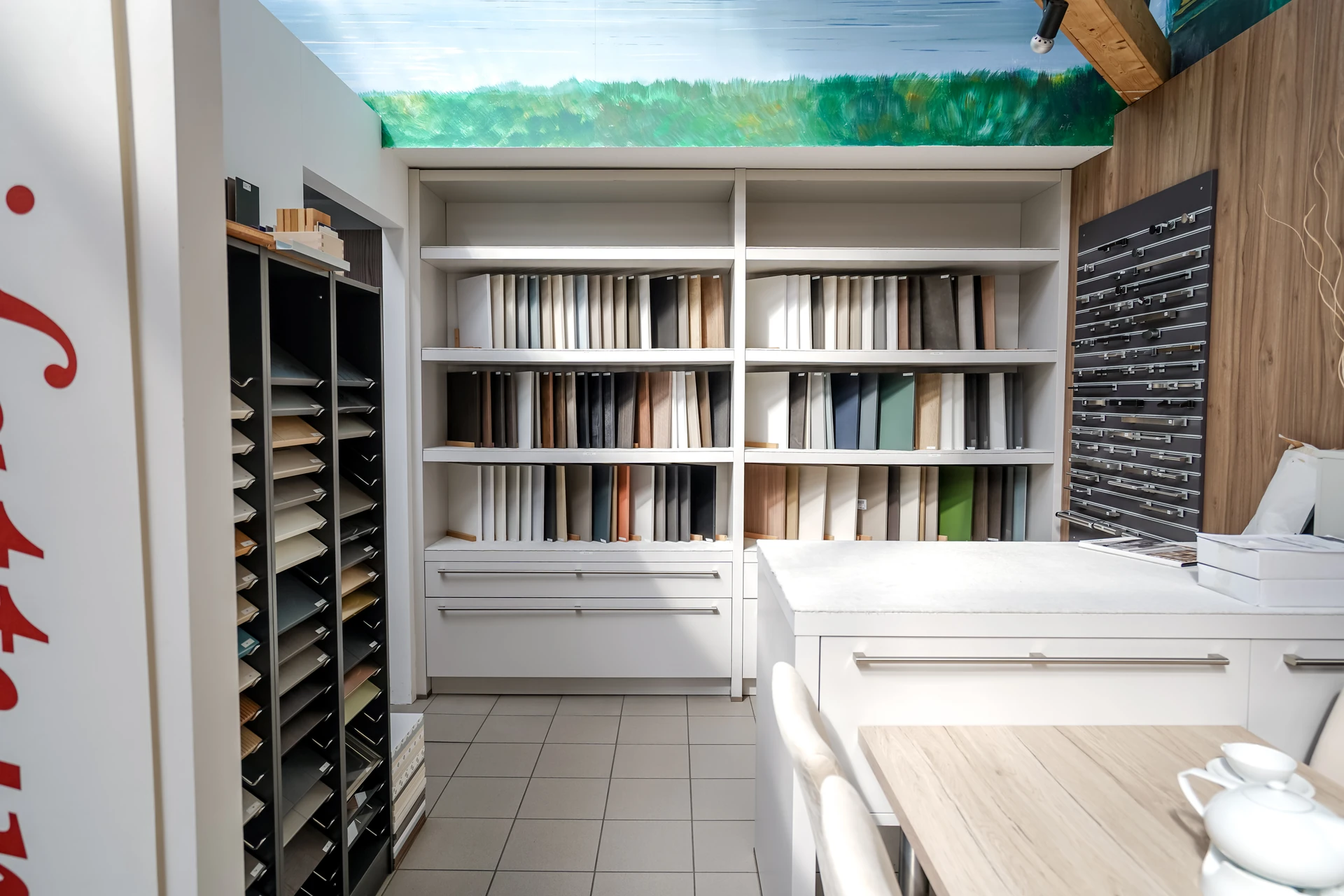 Regalwand mit aufrecht stehenden Dekor- und Stoffmusterbüchern unter bemalter Himmel-Wiesen-Decke, rechts eine weiße Ausstellungstheke.